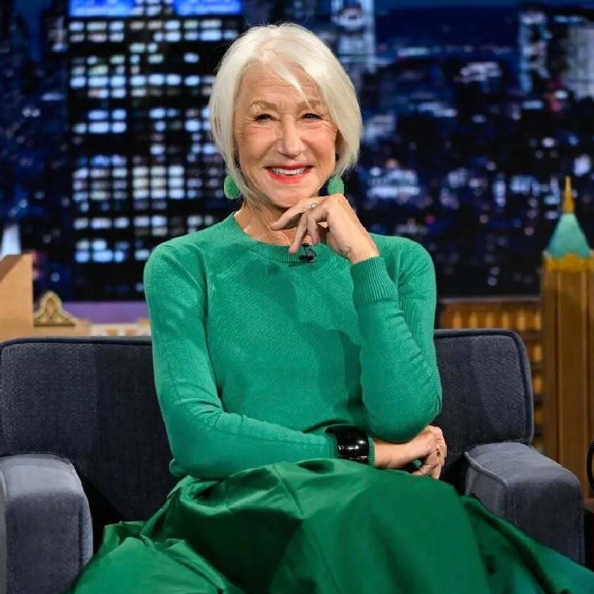 A woman with short white hair sits in a dark gray armchair on a talk show set, wearing a bright emerald green long-sleeve maxi dress and black heels, with a city skyline backdrop behind her.