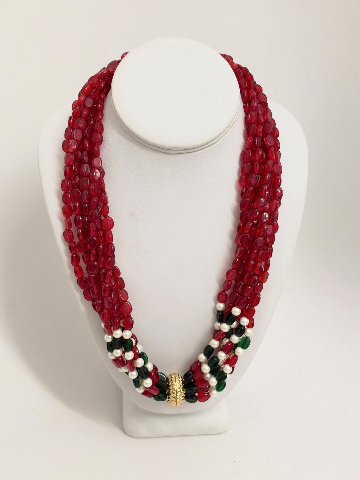 A multi-strand beaded necklace displayed on a white mannequin bust. The necklace features multiple strands of faceted red beads, with accent strands of white beads, dark green beads, and a textured gold clasp at the center.