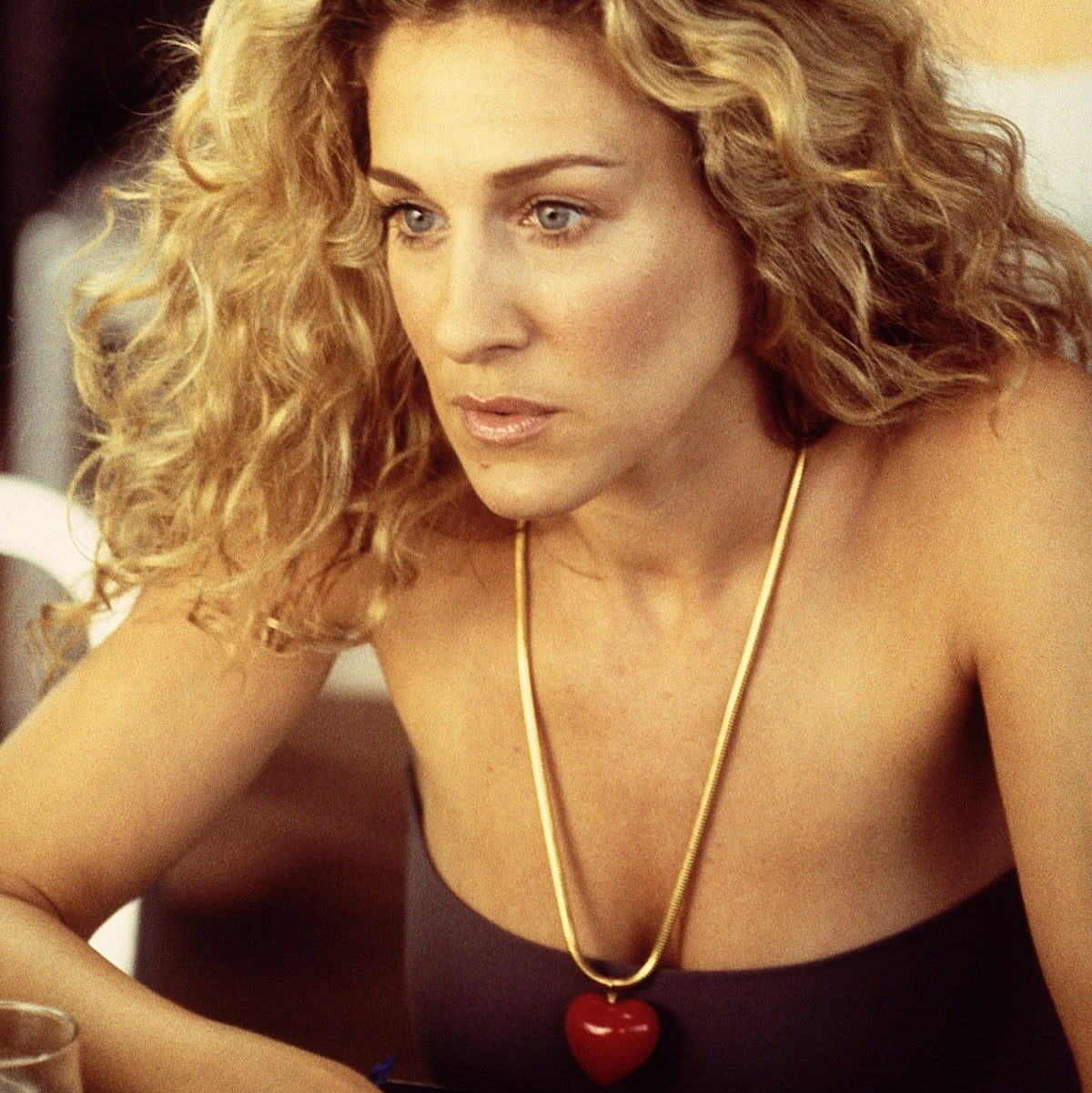 A close-up shot of a woman with curly blonde hair wearing a gold necklace with a red heart pendant, seated at a table with drinks.