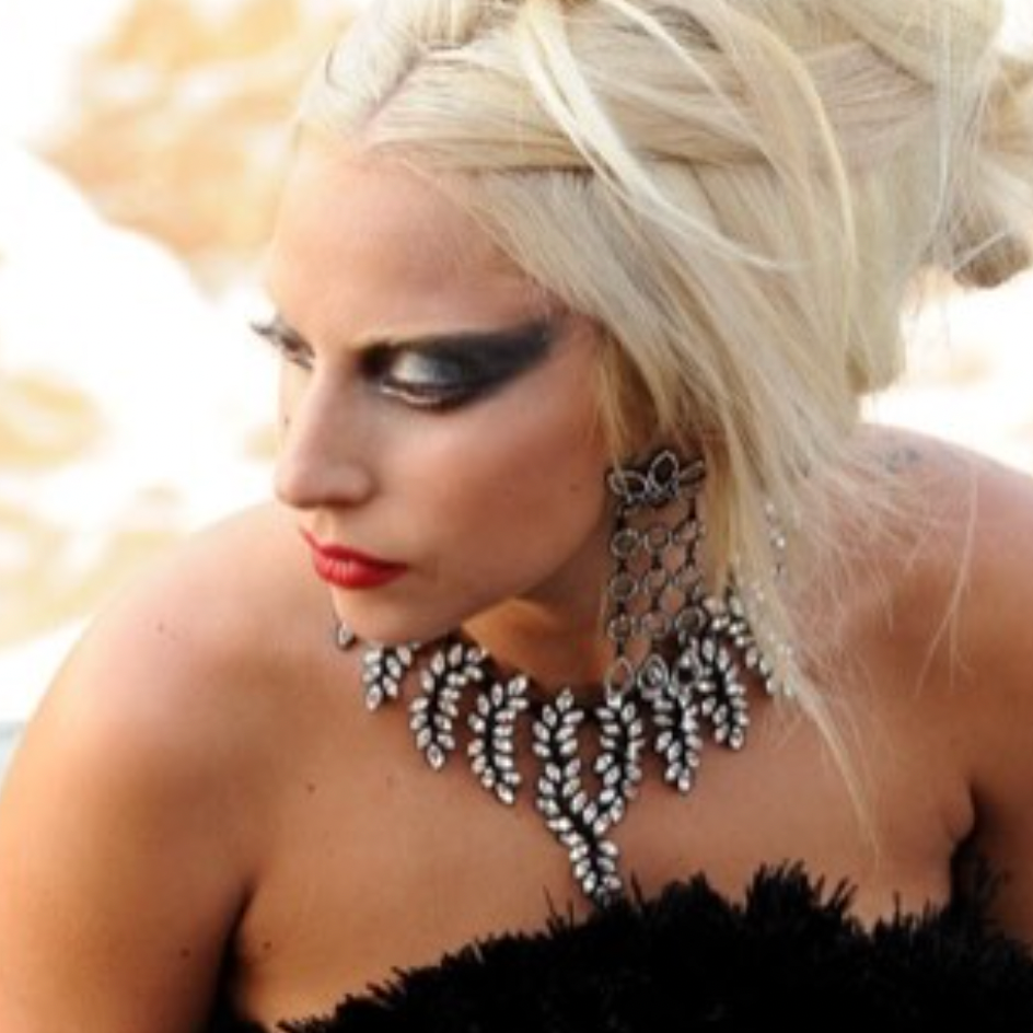 A close-up portrait of a woman with platinum blonde hair styled in an updo, wearing dramatic black eye makeup, red lipstick, a large silver-toned statement necklace, and matching dangling earrings, with a black feathered strapless top against a softly blurred light background.
